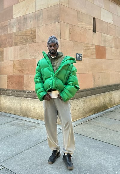 Chaqueta tipo puffa verde con un diseño recortado, superpuesta a una sudadera gris con capucha. Pantalones deportivos grises y calzado negro completan el conjunto.