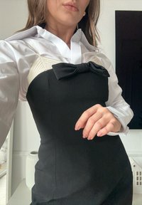 Femme portant une robe noire bustier avec un détail de nœud sur un chemisier blanc à manches longues, posant à l'intérieur avec un arrière-plan neutre.
