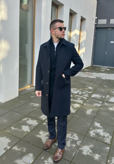 Abrigo de lana azul marino oscuro con cierre de botón, usado sobre un suéter negro. Combinado con pantalones oscuros y zapatos de hebilla de cuero marrón.