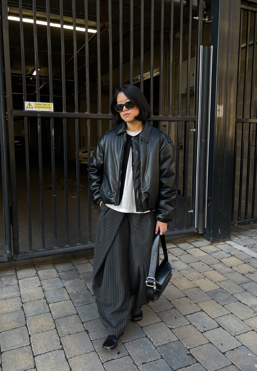 Personne portant une veste en cuir noire, une chemise blanche, un pantalon large à fines rayures, des lunettes de soleil noires et tenant un sac noir, debout près d'une porte métallique.