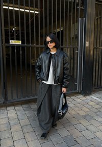 Personne portant une veste en cuir noire, une chemise blanche, un pantalon large à fines rayures, des lunettes de soleil noires et tenant un sac noir, debout près d'une porte métallique.