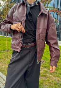 Casaco de couro bordeaux com gola, fecho de correr e bolsos dianteiros, combinado com uma camisa preta e calças que apresentam um detalhe em corrente.