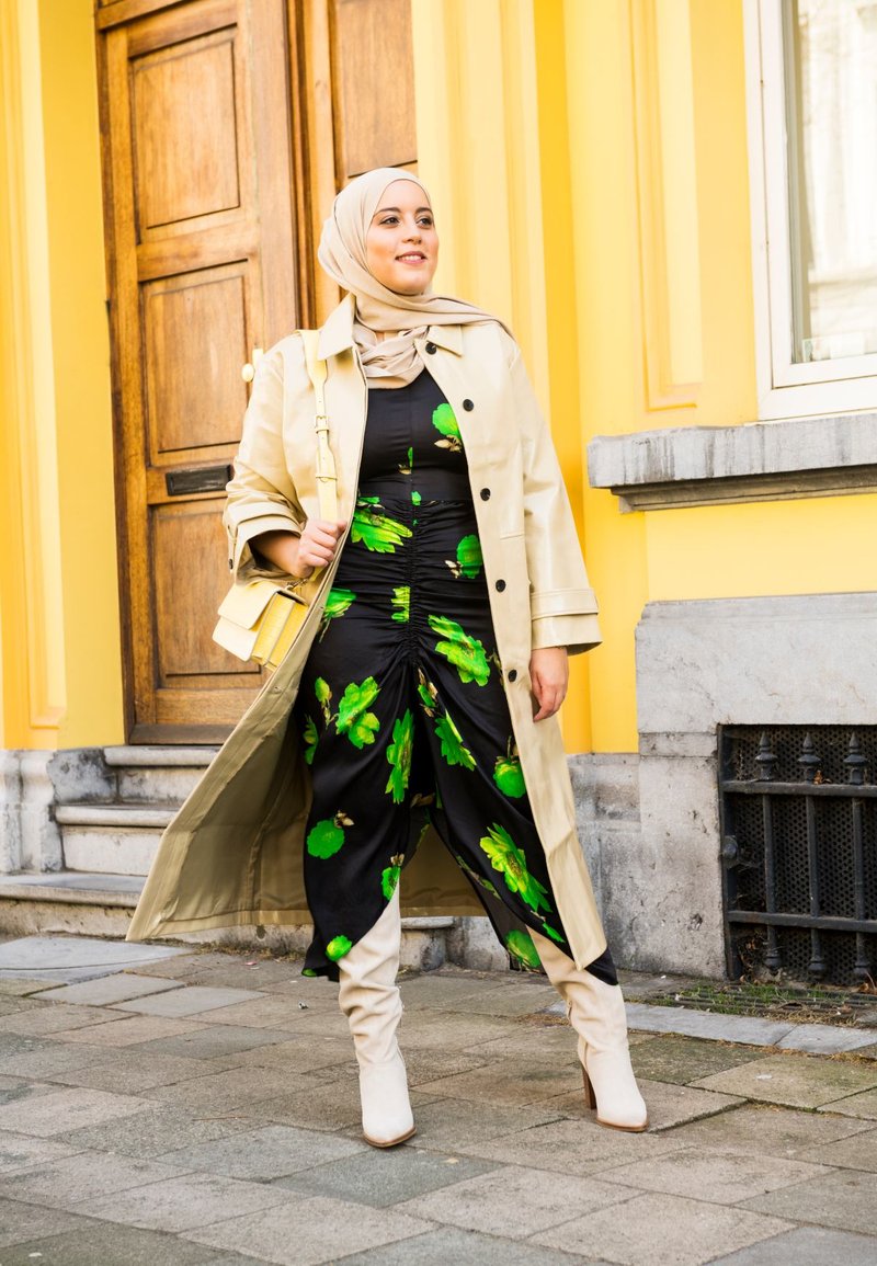Beige trench coat over a black dress with green floral patterns. Beige hijab and cream knee-high boots. Background features wooden door and steps.