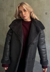 Μαύρο δερμάτινο παλτό με πλούσιο, σκούρο γκρι γούνινο γιακά και επένδυση. Βουργουνδίνο πουλόβερ από κάτω. Κουμπιά μπροστά στο παλτό.