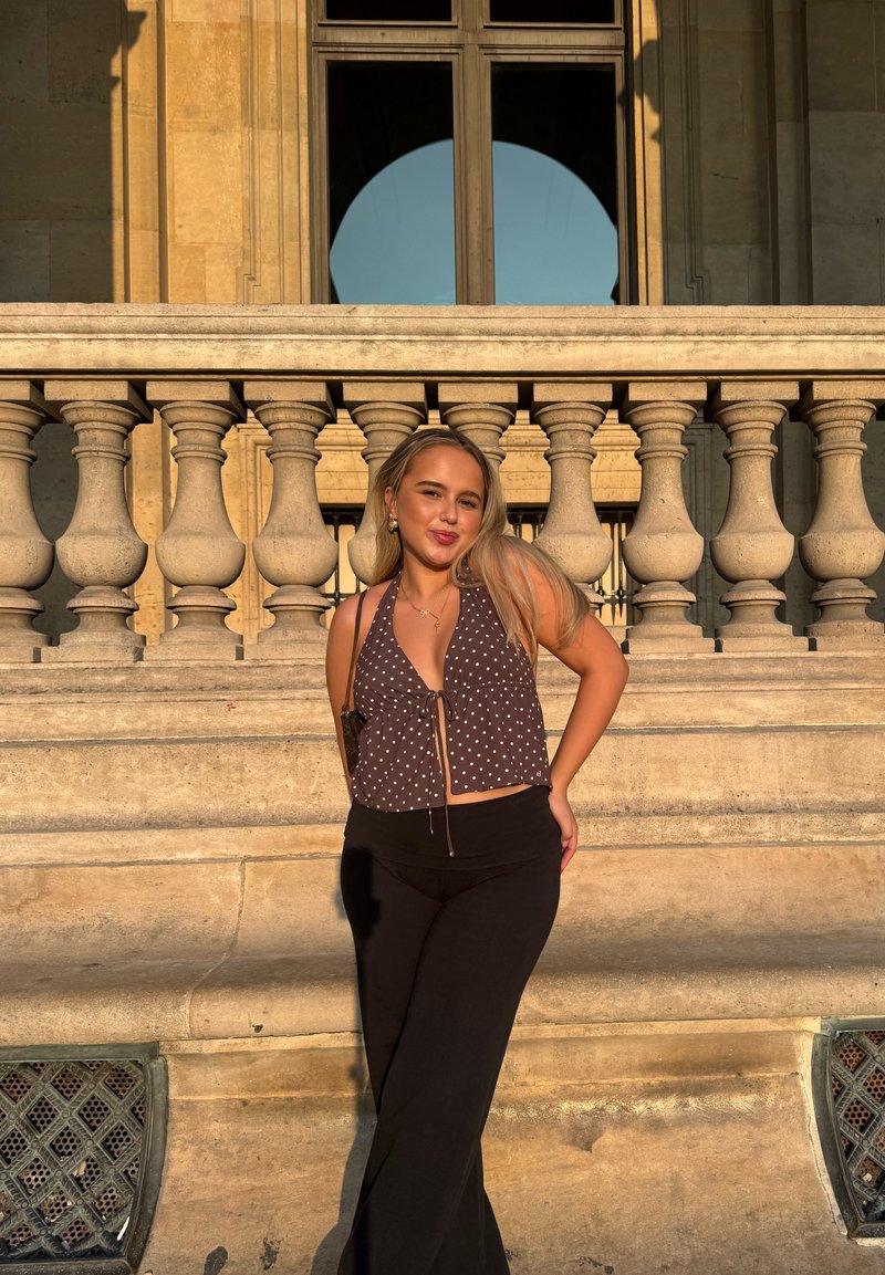 Braun-weiß gepunktetes Neckholder-Top mit Kordelzug, kombiniert mit schwarzen, ausgestellten Hosen. Im Hintergrund ist ein Steinbalustraden und ein Fenster zu sehen.