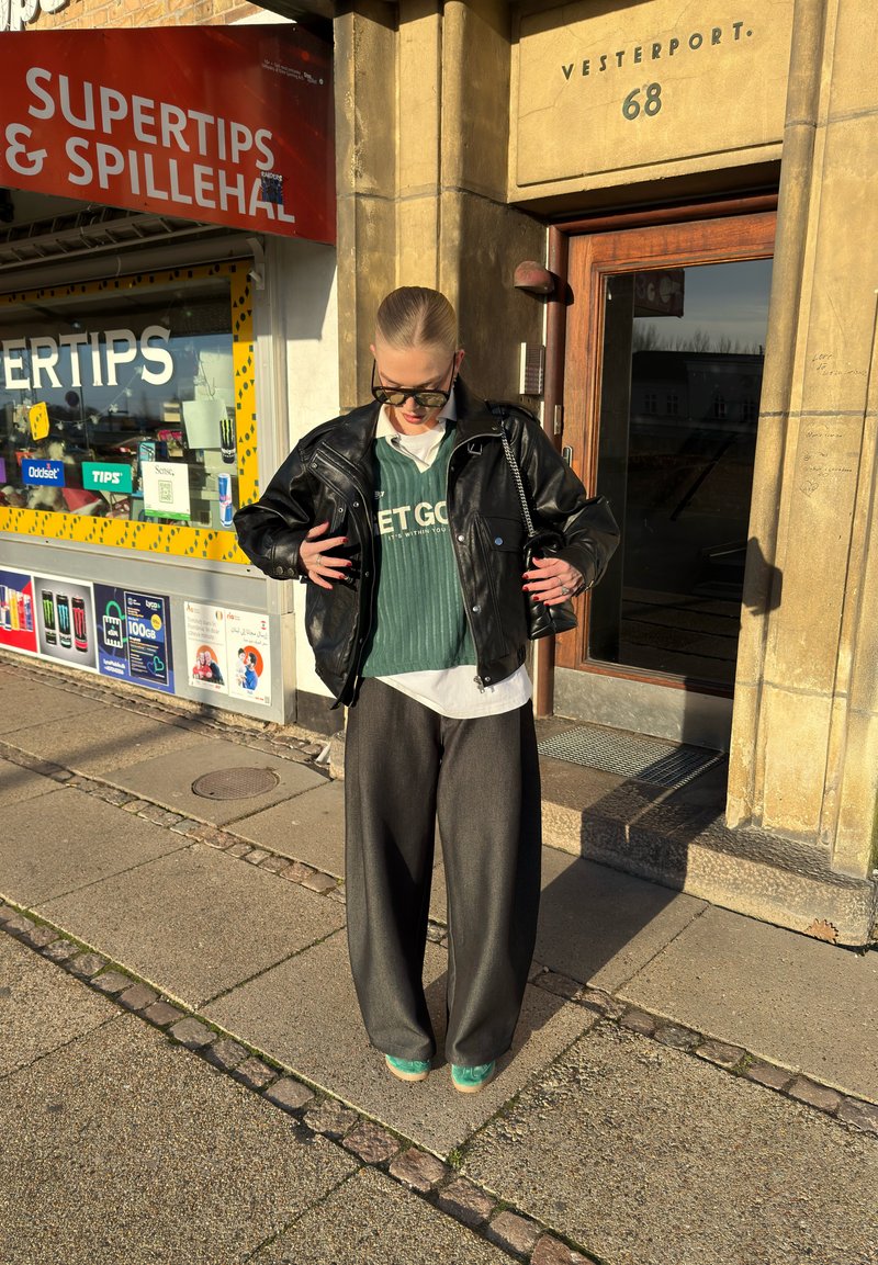 Veste en cuir noire, pull côtelé vert avec texte, chemise blanche à col, pantalon gris à jambes larges et chaussures turquoise. Debout en plein soleil.