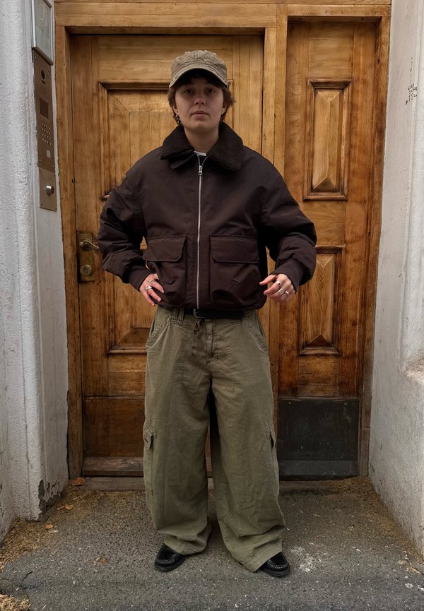 Giacca bomber marrone con collo in pile, due tasche frontali; pantaloni cargo larghi color verde oliva; scarpe nere; in piedi davanti a una porta di legno.