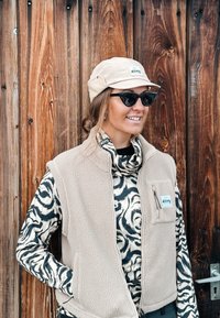 Femme souriante, portant un gilet en polaire beige, un col roulé noir et blanc à motifs, une casquette beige et des lunettes de soleil noires, debout devant un mur en bois.