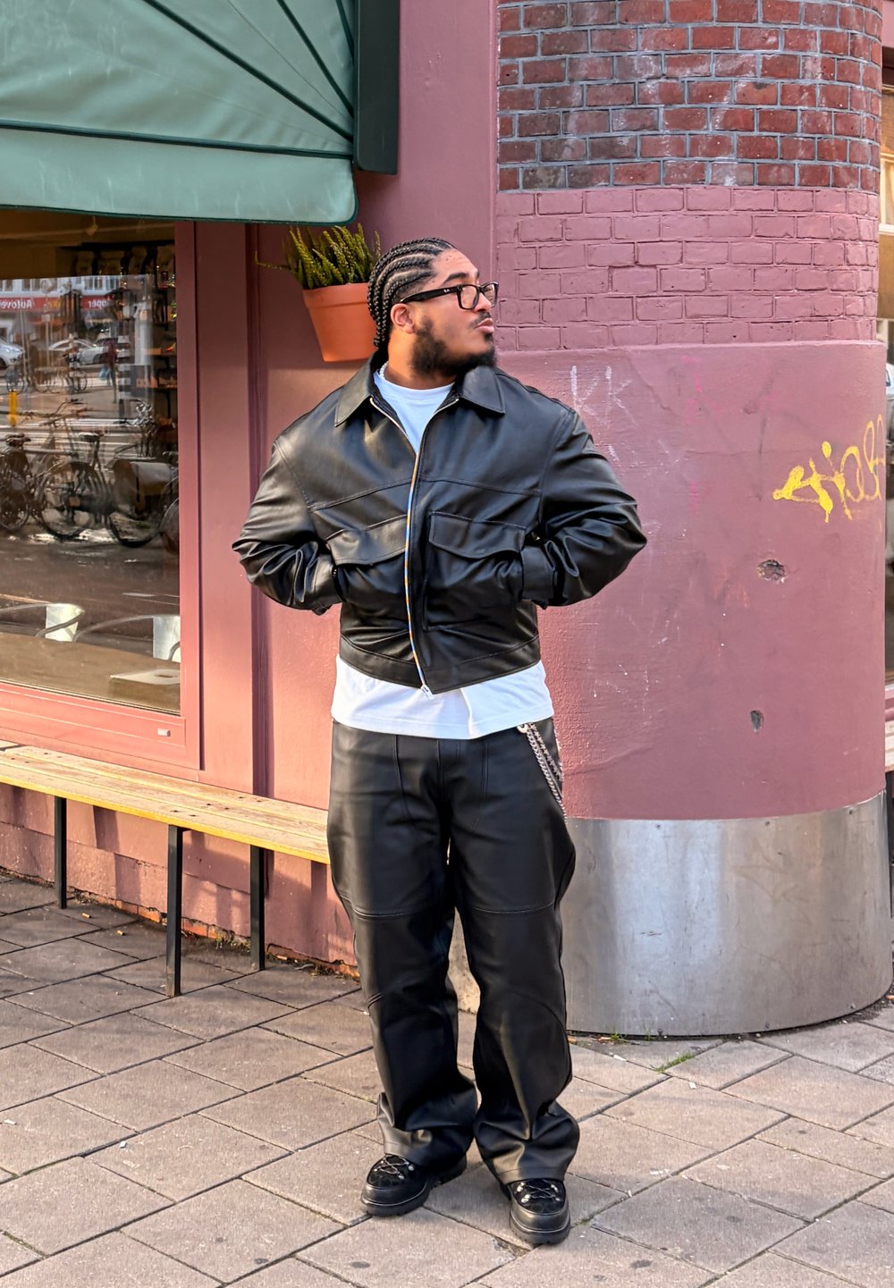 Homme aux cheveux tressés et portant des lunettes, vêtu d'une veste en cuir noire et d'un pantalon, se tient sur le trottoir à côté d'un bâtiment rose avec des graffitis.