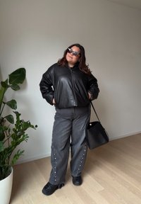 Mustast nahast bomberjakk tõmblukuga, hallid laiade säärega teksapüksid hõbedaste naeltega, mustad jalanõud ja päikeseprillid. Siseruumides roheluse taustal.