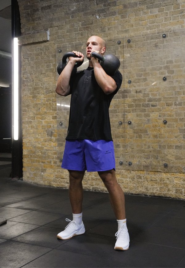 Man in zwart shirt en blauwe korte broek tilt twee kettlebells op schouderhoogte in een sportschool met een bakstenen muur op de achtergrond.