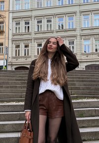 Langes braunes Haar, weißer grob gestrickter Pullover, dunkelbrauner Mantel, braune Lederhosen und eine braune Handtasche, stehend auf Steinstufen in der Nähe von Gebäuden.