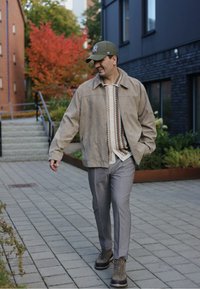 Beige suede jacket with a collared neckline, patterned shirt underneath, grey tailored trousers, and brown lace-up boots set in an outdoor environment.