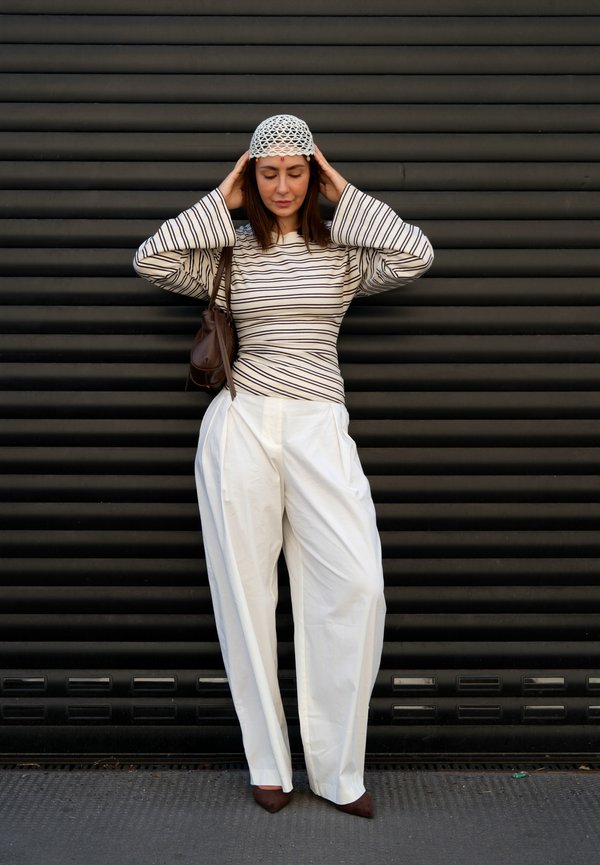 Femme ajustant un chapeau blanc au crochet, portant une chemise à manches longues rayée, un pantalon blanc large, des chaussures marron, et portant un sac à épaule marron, debout contre un mur noir.