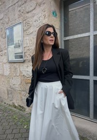 Black blazer over a black top, teamed with a white, voluminous skirt. Accessories include oversized black sunglasses and a circular pendant necklace.