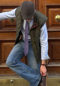 Gilet imbottito verde sopra una camicia azzurro chiaro, abbinato a una cravatta viola a fantasia e jeans blu. Scarpe marroni. Sfondo in pannelli di legno.