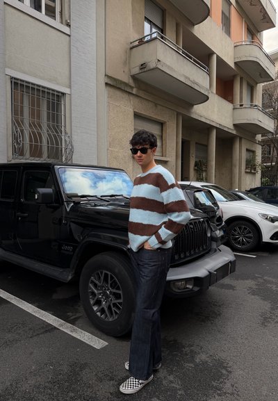 Joven con gafas de sol, suéter a rayas, jeans oscuros y zapatos slip-on a cuadros, de pie junto a un Jeep negro en un estacionamiento urbano.