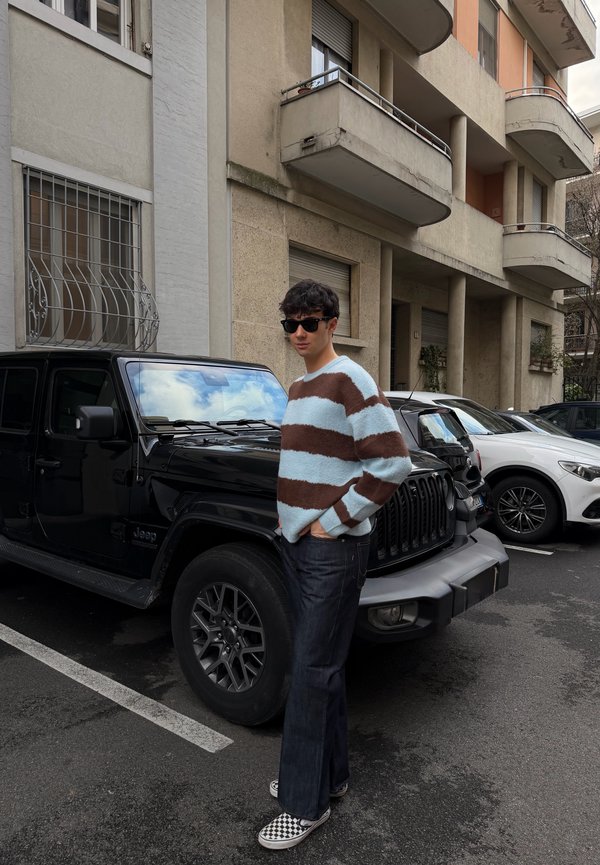 Jeune homme portant des lunettes de soleil, un pull rayé, un jean foncé et des chaussures à enfiler à carreaux, se tenant à côté d'un Jeep noir dans un parking urbain.
