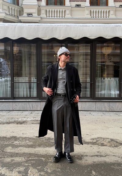 Hombre con abrigo negro largo, pantalones grises, polo gris, gorro gris, gafas de sol y zapatos negros, de pie frente a un edificio con grandes ventanas de vidrio.