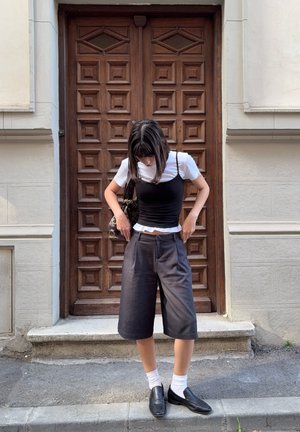 Schwarzes tailliertes Tanktop über einem weißen T-Shirt, kombiniert mit dunklen, locker sitzenden Shorts. Schwarze Slipper und weiße Socken. Strukturierte Holztür im Hintergrund.