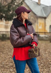 Jachetă scurtă din piele maro cu guler de blană neagră, pulover roșu tricotat, blugi albaștri și o geantă mică maro din blană cu lanț auriu.