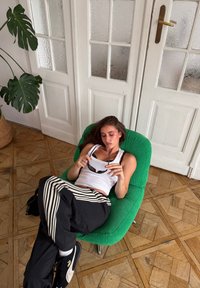 Poltrona lounge in velluto a coste verde con gambe cromate; persona con pantaloni a righe bianche e nere e top bianco, che tiene gli occhiali da sole, seduta in modo rilassato.