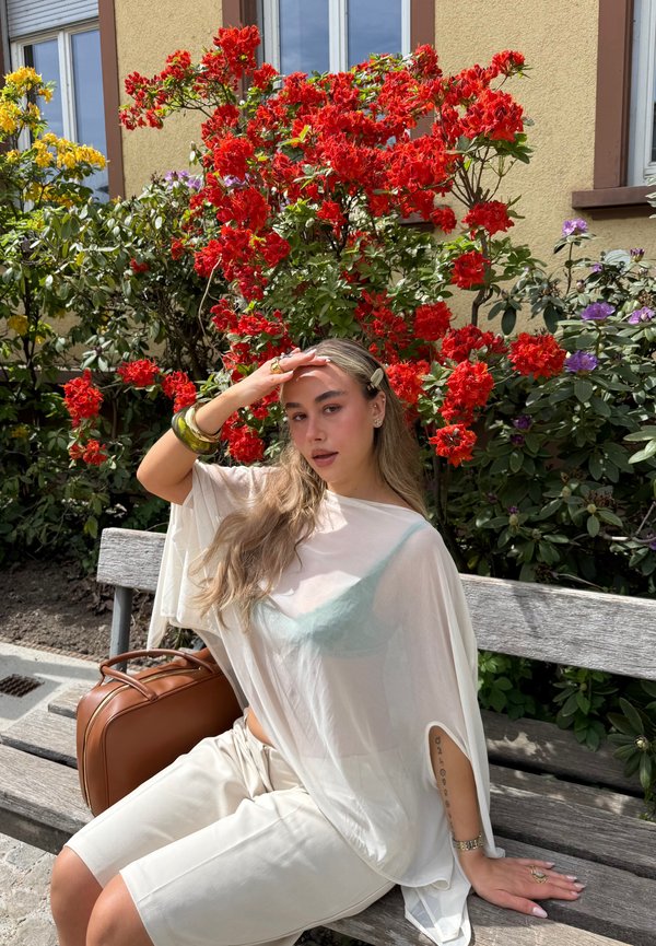 Giovane donna con capelli lunghi in abito bianco seduta su una panchina di legno, si scansa gli occhi, circondata da fiori rossi e gialli in fiore all’aperto.