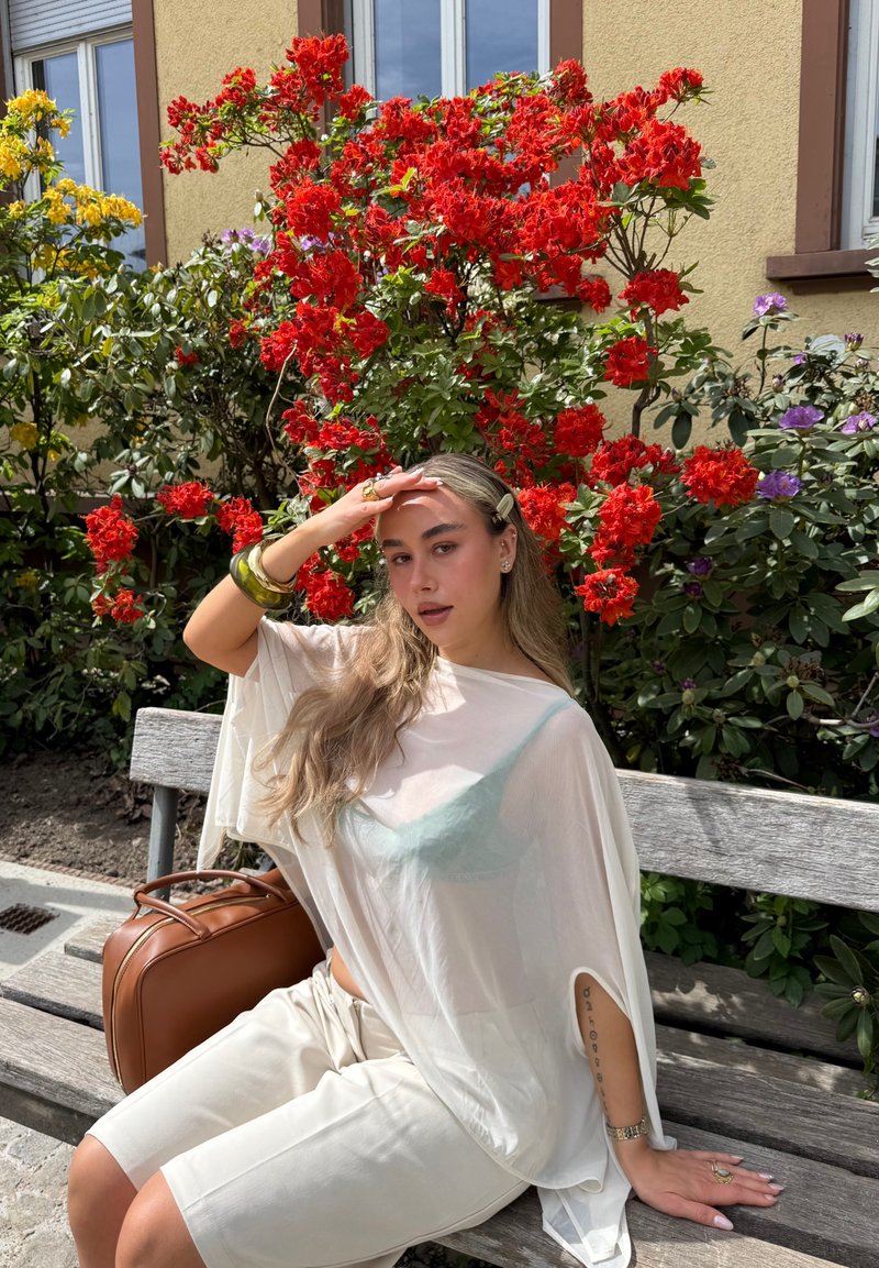 Jeune femme aux longs cheveux portant une tenue blanche, assise sur un banc en bois, se protégeant les yeux, entourée de fleurs rouges et jaunes en pleine floraison en extérieur.
