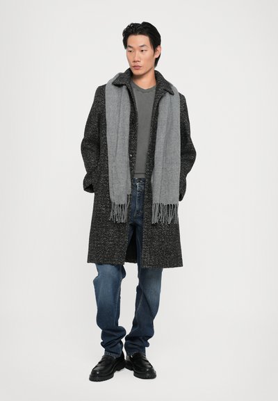 Abrigo largo negro y gris moteado con un cuello ancho, combinado con una bufanda gris, jeans azules y zapatos negros. Capas informales y texturizadas.