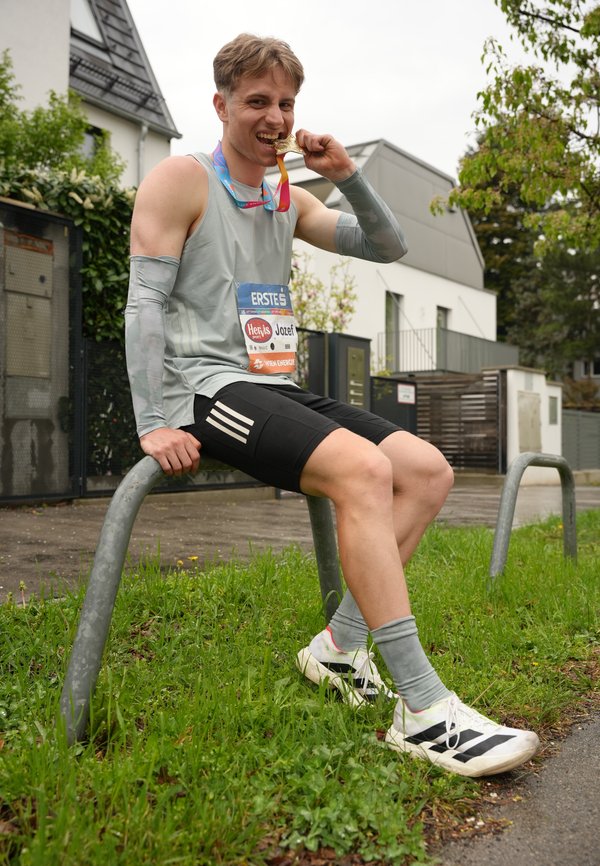 Jeune coureur masculin assis sur une rambarde en métal à l'extérieur, mordant une médaille d'or, portant un dossard et des vêtements de sport après une course.
