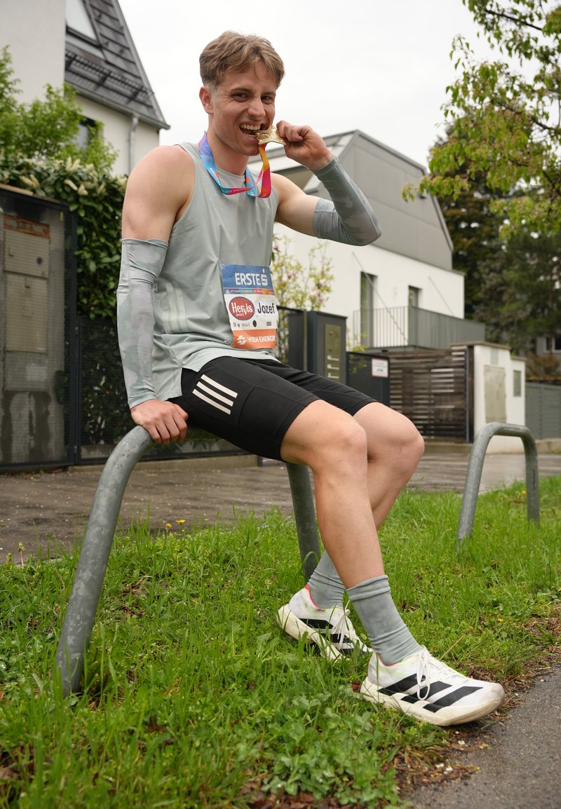 Jeune coureur masculin assis sur une rambarde en métal à l'extérieur, mordant une médaille d'or, portant un dossard et des vêtements de sport après une course.