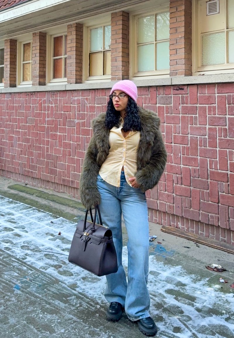 Femme portant un bonnet rose, des lunettes, une veste en fourrure, un t-shirt jaune, un jean bleu et des chaussures noires, tenant un sac à main noir sur un trottoir enneigé.