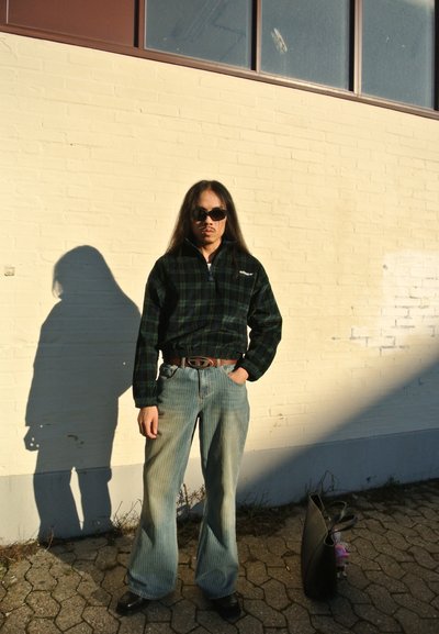 Chaqueta de cuadros negra y verde con cremallera, jeans de pierna ancha a rayas azul claro, cinturón negro y gafas de sol negras de gran tamaño. Sombra proyectada en la pared.