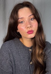 Grå ulltröja med rund halsringning och två knappar. Långt, vågigt mörkbrunt hår och naturlig sminkning med synliga fräknar och en septumring.