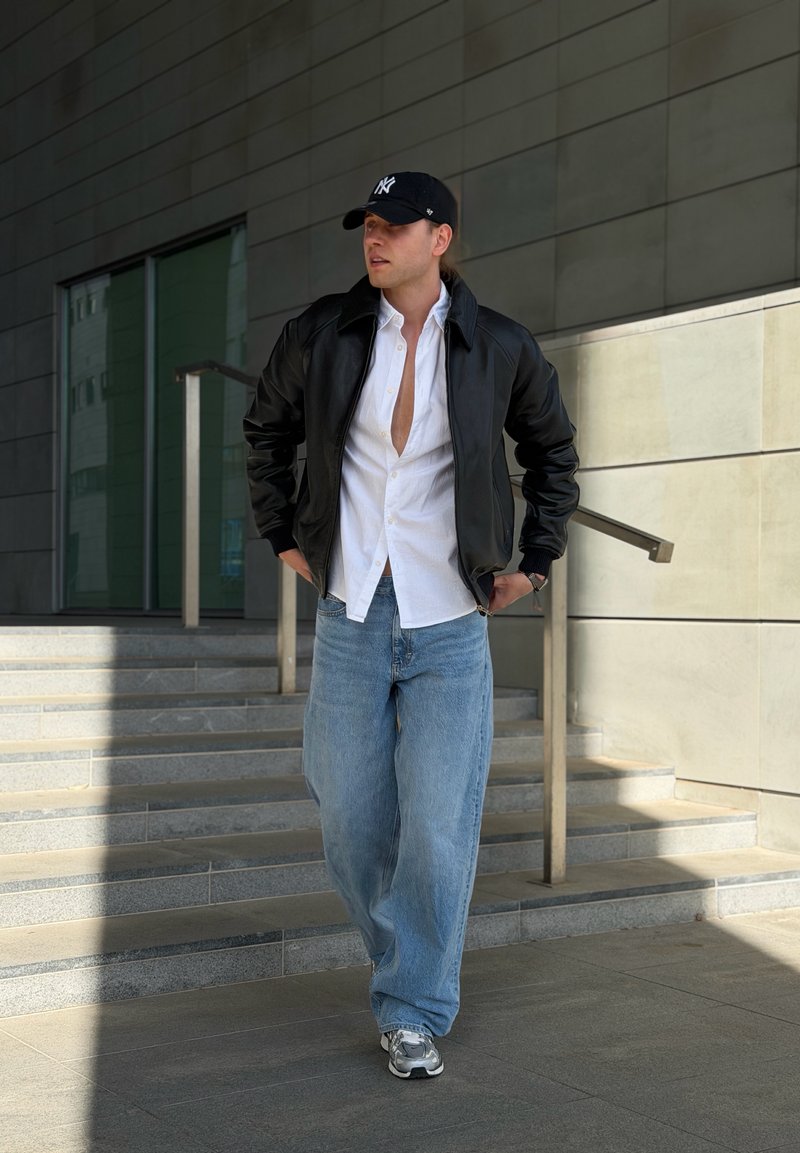 Jeune homme portant une veste noire, une chemise blanche ouverte, un jean bleu, des baskets grises et une casquette noire, descendant des escaliers à côté d’un bâtiment moderne.