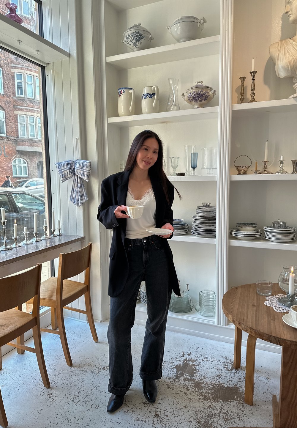 Blazer noir porté sur un haut blanc orné de dentelle, associé à un jean foncé. Tenant une tasse blanche avec sa soucoupe, entouré d'ustensiles de cuisine décoratifs.