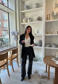 Blazer noir porté sur un haut blanc orné de dentelle, associé à un jean foncé. Tenant une tasse blanche avec sa soucoupe, entouré d'ustensiles de cuisine décoratifs.