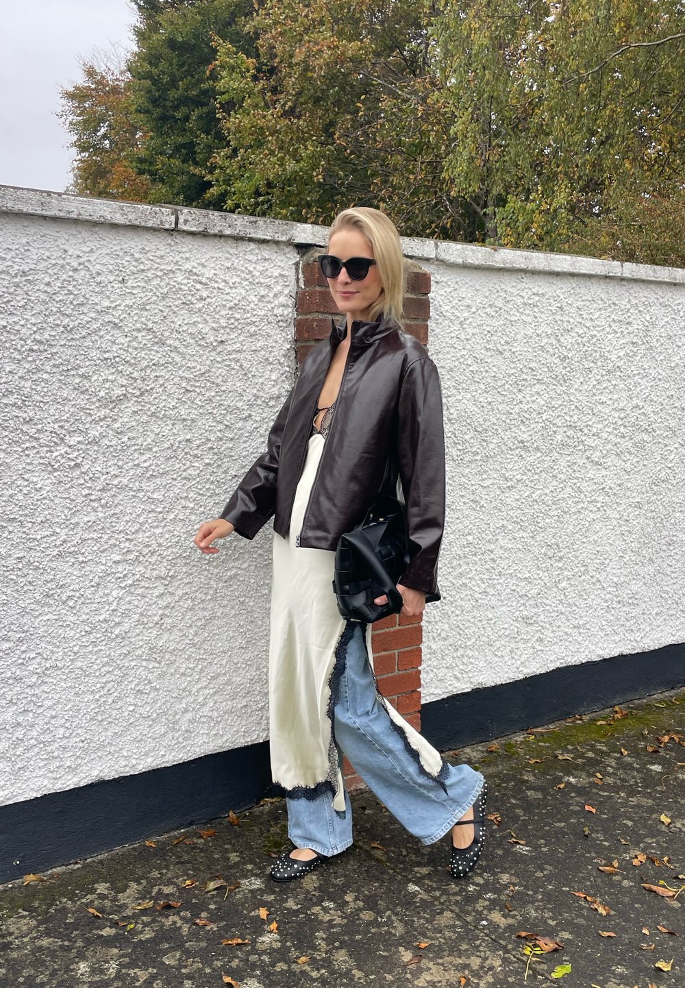 Jaqueta de couro castanho, vestido de cetim branco com detalhes em renda, calças largas de denim azul-claro, sapatos planos pretos com tachas e uma bolsa preta.