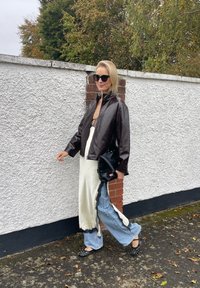 Jaqueta de couro castanho, vestido de cetim branco com detalhes em renda, calças largas de denim azul-claro, sapatos planos pretos com tachas e uma bolsa preta.