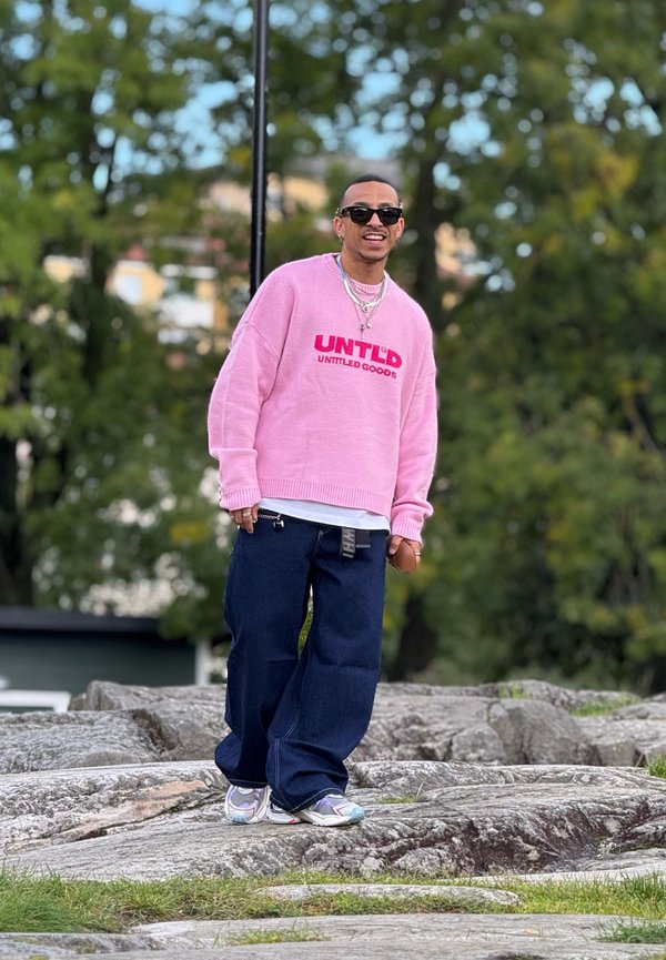 Sweater oversized rosa com o logótipo "UNTLD", combinado com calças de ganga largas azul escuro e ténis multicoloridos, em pé sobre um terreno rochoso.