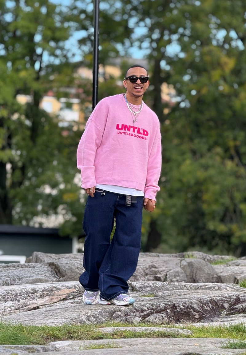 Sweater oversized rosa com o logótipo "UNTLD", combinado com calças de ganga largas azul escuro e ténis multicoloridos, em pé sobre um terreno rochoso.