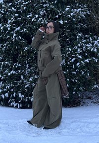 Trench-coat vert olive avec un col haut, des détails à boutons et une ceinture. Associé à un pantalon marron ample et texturé, posé sur de la neige.