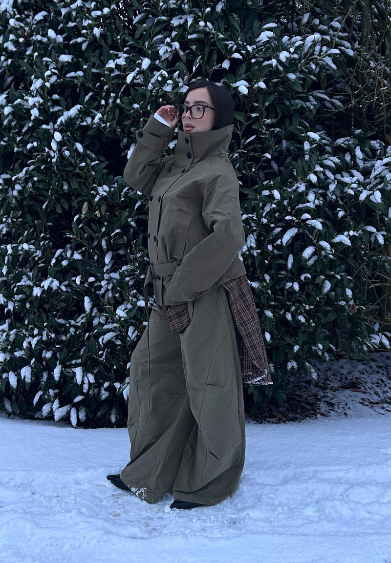 Trench-coat vert olive avec un col montant, des détails à boutons et une ceinture. Associé à un pantalon marron ample et texturé, sur fond de neige.