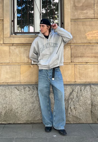 Sudadera gris con cremallera y texto "GAP ATHLETIC", jeans de mezclilla azul de corte holgado y zapatos negros. De pie frente a una pared de piedra texturizada.