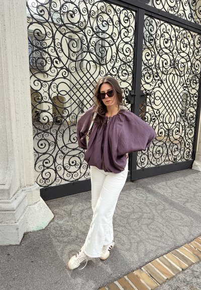 Mujer con blusa morada, pantalones blancos y gafas de sol, de pie con las manos en las caderas frente a una elaborada puerta de hierro forjado negra.