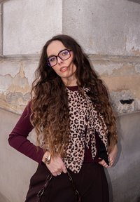 Lapu print šalle, kas valkāta virs bordo garām piedurknēm; teksturēti mati; bronzas rokassprādze; brilles ar violeta nokrāsu; brūnas bikses.