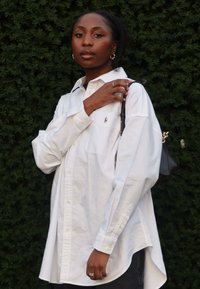 Biała koszula na guziki z jedną kieszenią i małym logo, zestawiona z ciemnymi dżinsami i czarną torbą. Gładka faktura, luźny krój.