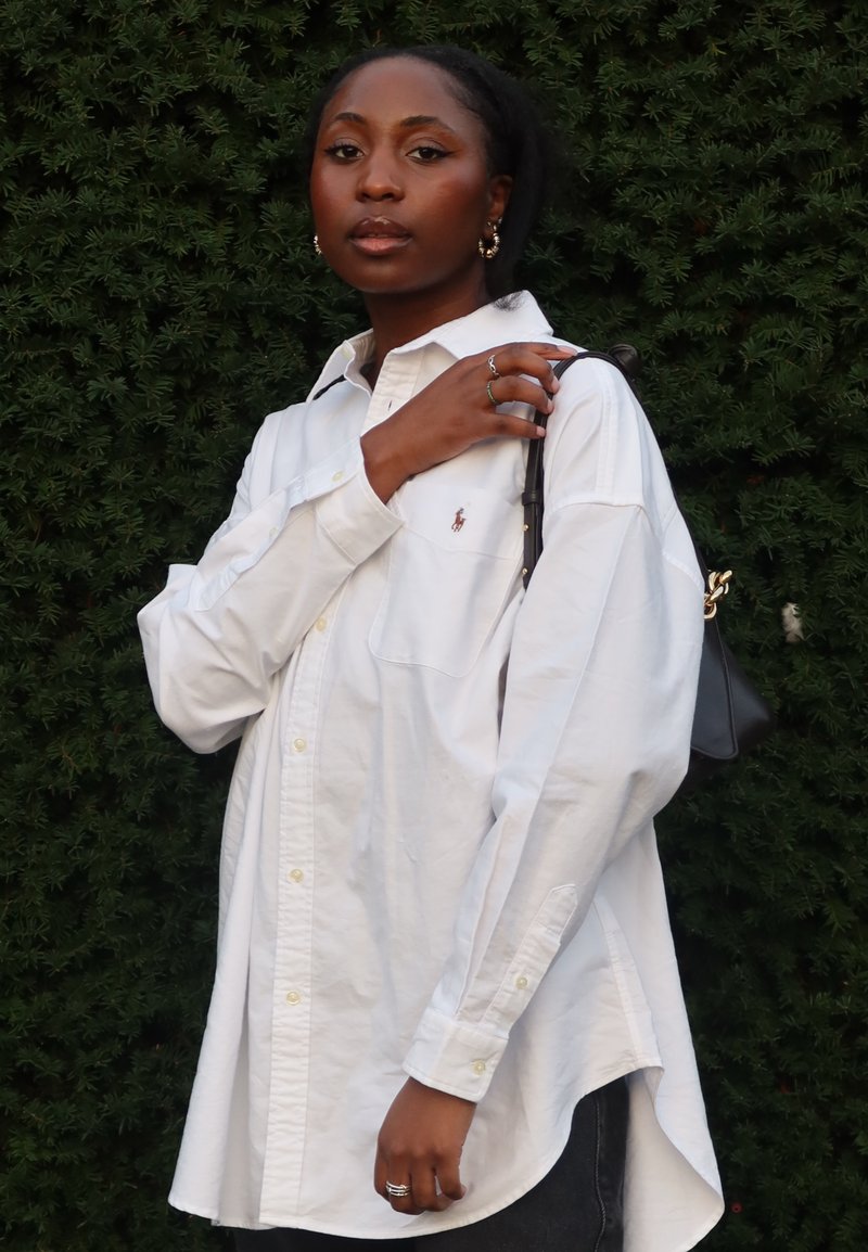 Chemise blanche boutonnée avec une poche unique et un petit logo, assortie à un jean foncé et un sac noir. Texture lisse, coupe décontractée.