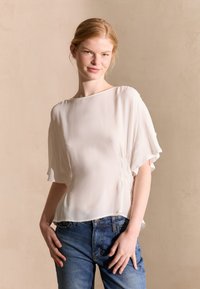 Blusa beige chiaro in tessuto fluido e trasparente con scollo a barca e maniche a sbuffo larghe. Presenta dettagli arricciati sui lati e una texture morbida.