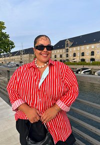 Red and white striped button-up shirt with a light blue accent, paired with black shorts and a small black handbag, worn outdoors by a smiling individual.